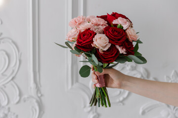 A hand holds a romantic mixed bouquet of deep red roses, blush roses, and pink carnations against a classic white wall with stucco. Features ample copy space on the left