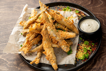 Golden brown deep-fried fish served on parchment paper with creamy dipping sauce, herbs, and spices. Rustic wooden background. Appetizing food presentation for culinary use.