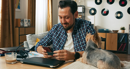 A young, delighted, handsome man wearing a shirt sits at the table with a cheerful small puppy, smiling while holding a credit card and making an online purchase from home.
