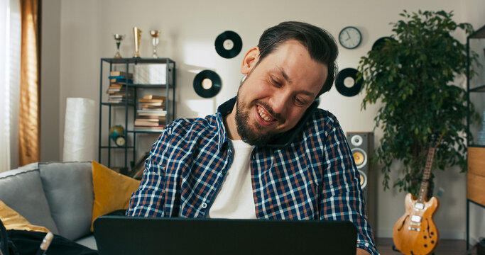 A bearded young male office worker enjoys a call with friends or colleagues, smiling joyfully as he learns about a weekend party, celebrating Friday and the end of the week.