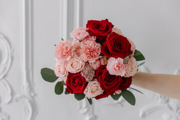 A hand holds a romantic mixed bouquet of deep red roses, blush roses, and pink carnations against a classic white wall with stucco. Features ample copy space on the left