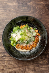 Gourmet shrimp dish with mashed potatoes, fresh greens, Parmesan cheese, and microgreens served on a black plate over wooden table. Modern restaurant-style food presentation.