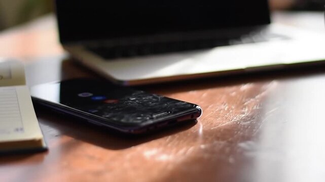 Close-up view of a damaged smartphone with a severely cracked and shattered glass screen lying on a wooden business desk next to a laptop