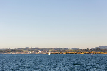 Obraz premium A lone sailboat sails on the blue water of the Bay of Santander on a sunny day, with the coastline and mountains visible in the distance.