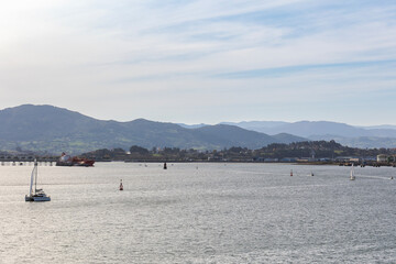 A scenic view of the Bay of Santander, Spain, on a sunny day. Sailboats and a cargo ship are on the water, with the city's port and mountains in the background.