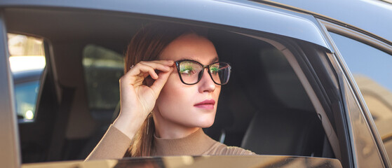 Young, confident businesswoman wearing glasses looks out of car window with excitement