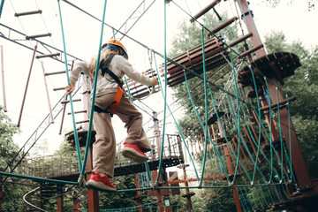 Young child wearing safety harness and helmet skillfully navigates a challenging high rope course. Surrounded by trees, this outdoor adventure highlights courage and balance.