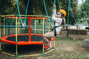 Young child wearing safety gear makes their way across a challenging ropes course in an adventure park. The image captures focus and determination during this outdoor activity amid lush surroundings.