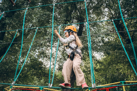 Young child wearing safety gear makes their way across a challenging ropes course in an adventure park. The image captures focus and determination during this outdoor activity amid lush surroundings.
