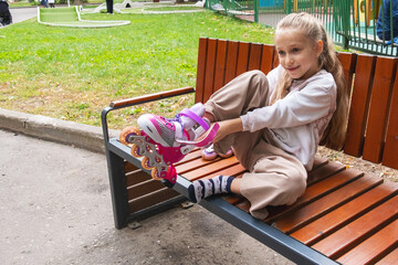 Young girl happily putting on pink roller skates while sitting on a park bench. The outdoor setting adds to her excitement as she prepares to enjoy a skating session.