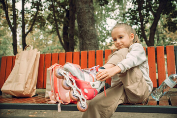 Young girl sitting on park bench, lacing up roller skates beside a bright pink backpack. The setting suggests outdoor playtime, emphasizing a sense of fun and activity in nature.