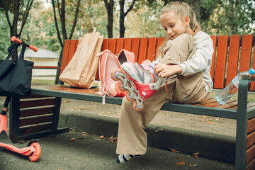 Child sitting on a park bench, focused on putting on roller skates. A backpack beside them hints at an adventurous day. Leaves surround the area, adding a touch of nature.