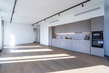 A sleek kitchen is featured in a spacious living area, illuminated by sunlight pouring through large windows. The minimalist design includes contemporary cabinets and open space
