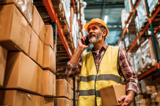 A warehouse worker in a reflective vest and hard hat stands among stacked boxes, talking on a mobile phone while managing inventory in a bustling storage space