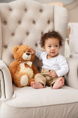 Cute baby sitting on soft armchair with teddy bear, looking curious and happy. Warm home interior, childhood, comfort, and innocence captured in cozy natural light.