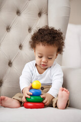 Obraz premium Cute baby sitting on chair playing with colorful stacking toy. Toddler developing fine motor skills, learning colors, concentration and coordination in cozy home environment.