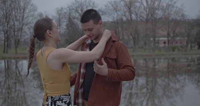 A loving couple shares an affectionate and playful moment together by a tranquil lake in a park setting. They are laughing and adjusting clothing, showcasing their close bond and happiness outdoors.