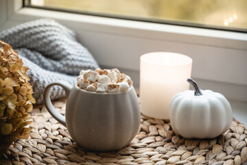 Autumn warming chocolate or coffee with marshmallows, pumpkins and candle on window sill. Autumn season beverage. Close up.