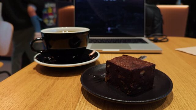 A delicious coffee, a rich brownie, and a laptop are placed on a wooden table in a cozy cafe setting