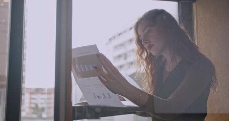 Young business woman reviewing charts and graphs in a modern office space. She is wearing a headset, suggesting she is on a call or listening to music. - Powered by Adobe