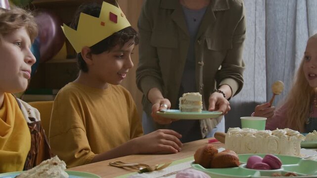 Boy in paper crown hungrily eating piece of cake with his hands, while his friends communicating at table during children birthday celebration