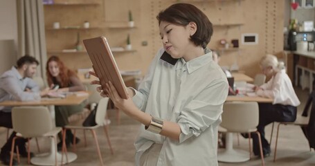 A young, Asian businesswoman multitasking in a busy startup office environment. She holds a tablet and talks on her phone while colleagues collaborate in the background. - Powered by Adobe
