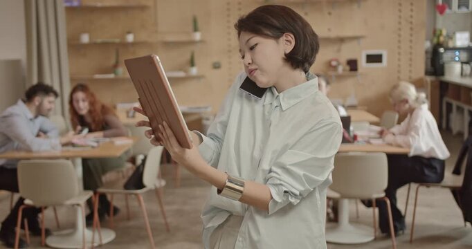 A young, Asian businesswoman multitasking in a busy startup office environment. She holds a tablet and talks on her phone while colleagues collaborate in the background. - Powered by Adobe