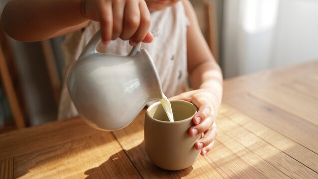 Young child's hands pouring fresh milk from a ceramic jug into a matching cup on a sunlit wooden kitchen table, showing independence and a simple morning routine