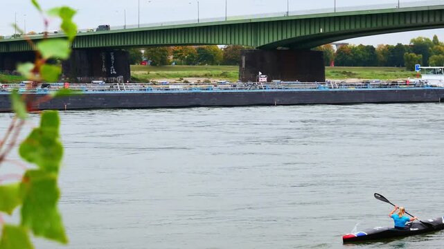 Rowing Woman in Kayak under Green Bridge over River with Cargo Barge Passing Amidst Autumn Trees and Urban Riverside Landscape on Overcast Day
