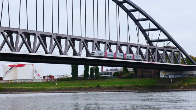 Stadtbahn Train Crossing Deutzer Bridge over Rhine River in Cologne Germany with Cargo Barge Sailing Beneath Amid Industrial Tanks Graffiti Walls and Urban Landscape on Overcast Day