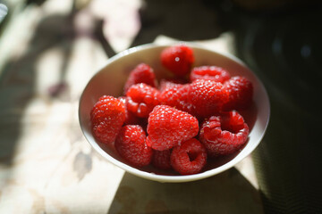 A bowl brimming with ripe raspberries catches the sunlight, creating a vibrant contrast against the table. The natural light enhances the rich red color of the fruit.