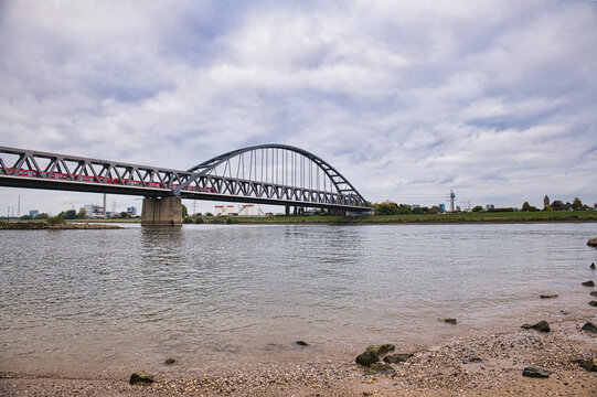 Modern steel arch bridge with red commuter train crossing river on cloudy day, transportation and urban infrastructure concept