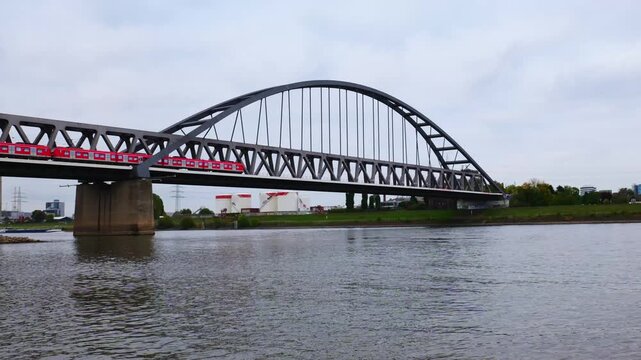 Red Stadtbahn Train Crossing Deutzer Bridge over Rhine River in Cologne Germany with Industrial Tanks and Urban Riverside Landscape on Overcast Day