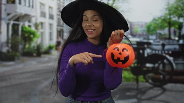 Latin woman wearing purple shirt and black witch hat pointing finger to orange pumpkin bucket on crowded street; joy. - Powered by Adobe