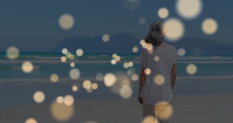 Standing senior woman gazing toward horizon at sandy beach, with golden bokeh lights, copy space