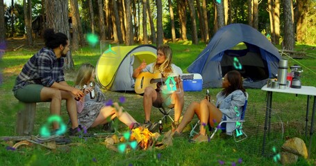 Fototapeta premium Sitting around campfire four friends chatting in forest clearing, with acoustic guitar, tents
