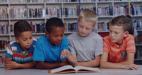 Leaning four school age boys reading open hardcover book in library reading area, with bookshelves