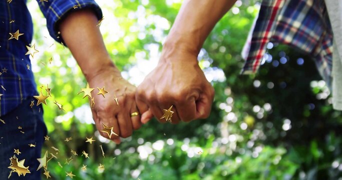 Linking senior couple in plaid shirts hooking pinkies in garden with trees, with gold wedding bands