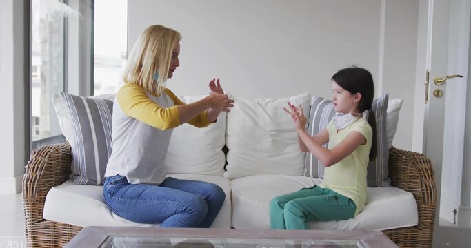 Practicing mother and daughter signing on wicker sofa near large window at home, wearing masks