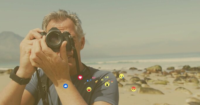 Holding DSLR camera to eye, photographer capturing ocean waves at sandy shoreline, floating emojis