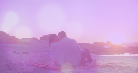 Sitting couple sharing light-colored blanket on pink fringed beach blanket at sunset, pastel light