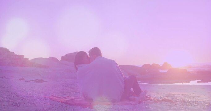 Sitting couple sharing light-colored blanket on pink fringed beach blanket at sunset, pastel light