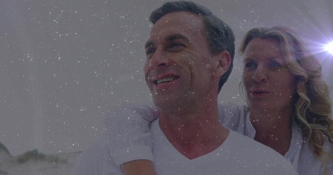 Laughing couple wearing white T-shirt and blouse posing piggyback on sandy beach, with lens flare