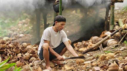 Indonesian farmer opening coconut with machete for making dried copra, smoke drying rack in...