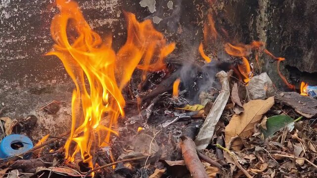 Bright orange flames consume a pile of trash, wooden twigs (ranting kayu), and dry leaves (dedaunan kering), creating smoke and intense heat.
