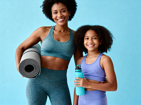 Happy Black mother and daughter in athletic wear smiling with yoga mat and water bottle against a blue background, promoting family fitness and wellness. - Powered by Adobe