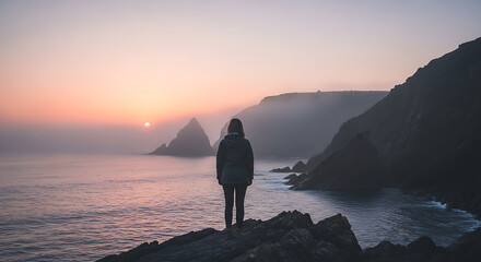 Silhouette of a person standing on rocky cliff overlooking calm ocean sunset with distant mountains