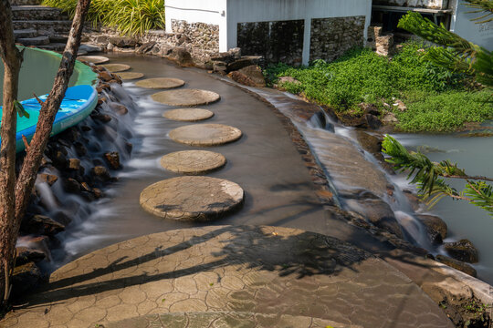 Round stone steps curve above the gentle waterfall, beside a turquoise paddle board at De'camp hill Ka-ang resort, Nakhon Nayok, Thailand.