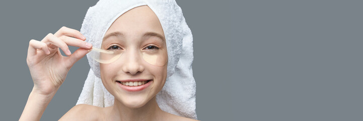 Young caucasian female applying eye patches smiling in towel headwrap on gray background.
