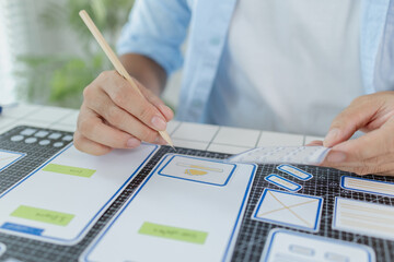 A designer is developing UX UI layouts, reviewing mobile app prototypes, and arranging interface elements on paper and board. He is focused on user experience and digital creativity.